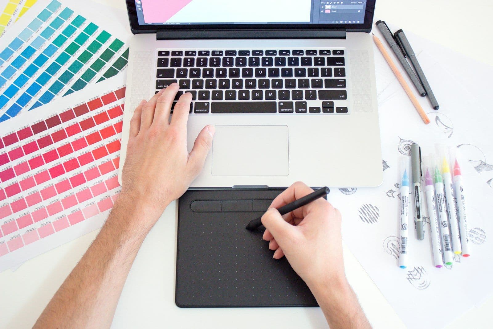 A designer's hand with colour swatches beside a laptop and a drawing tablet